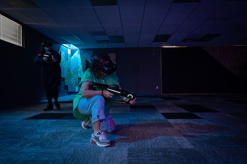 A player in a headset crouches with a blaster in a dark, blue-lit arena.