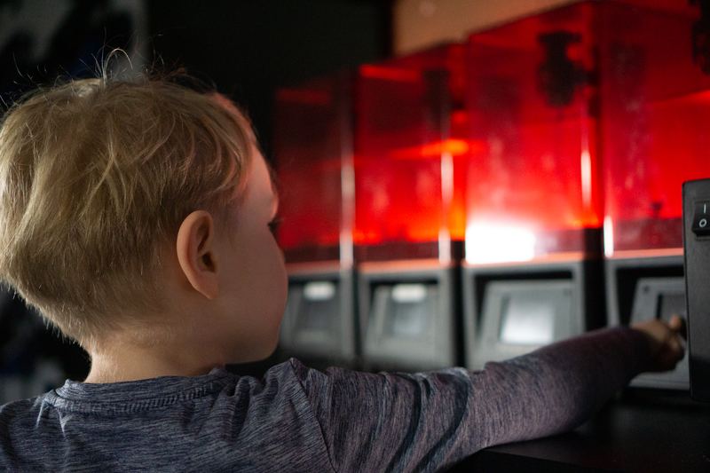 A young camper reaches toward a row of 3D printers glowing red.