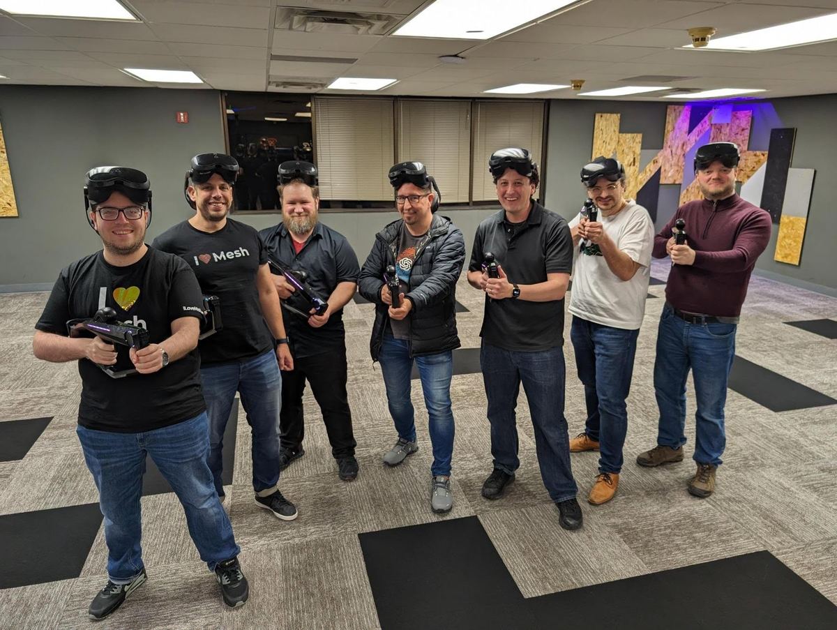 Seven coworkers pose with VR blasters in the arena, headsets pushed up on their heads.