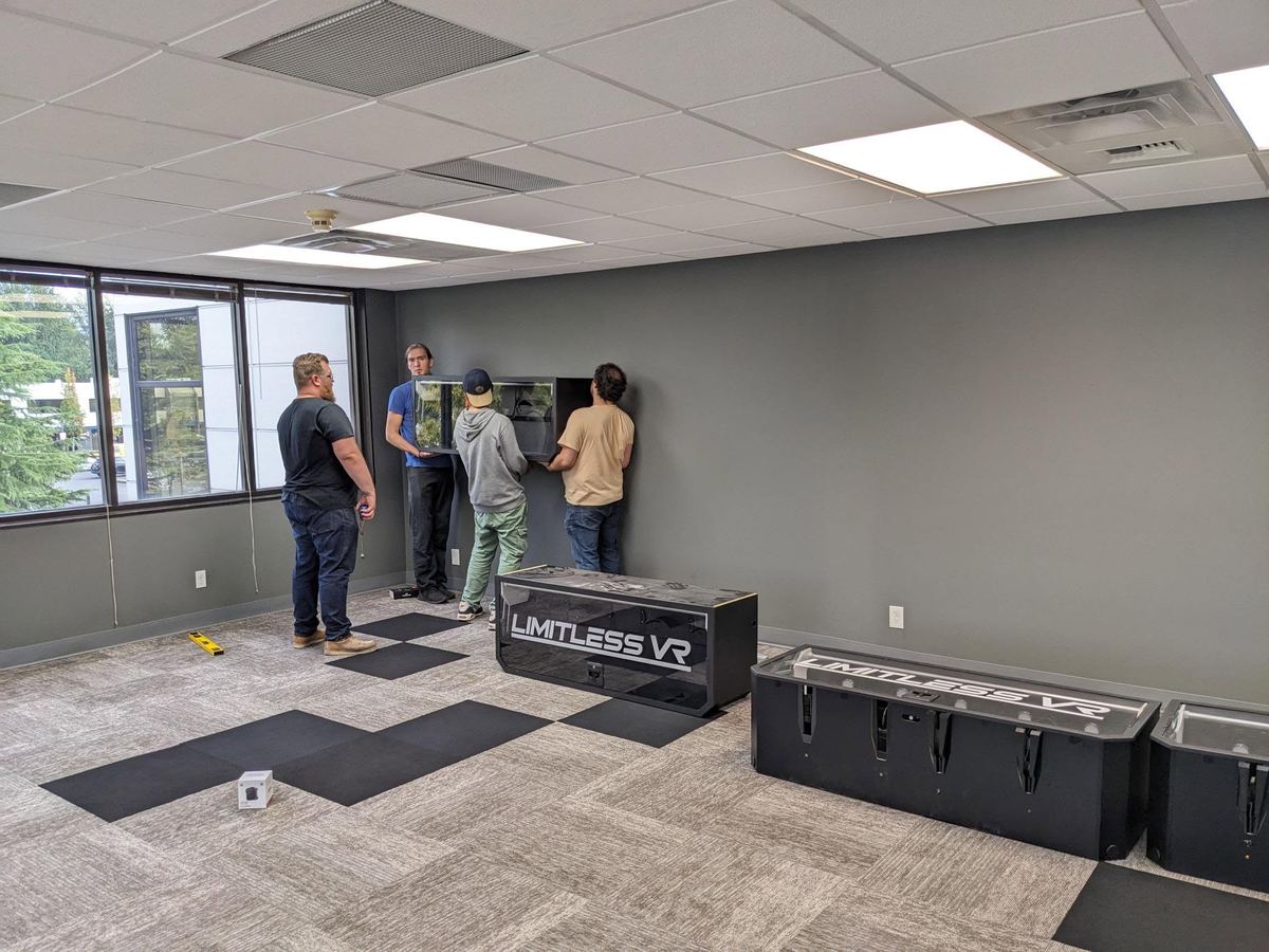A crew mounts racks on the wall of a bright, empty room, with LimitlessVR equipment cases on the carpet.