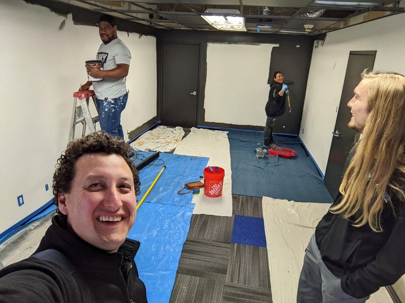 A grinning selfie in front of a crew painting the walls black, with tarps on the floor.