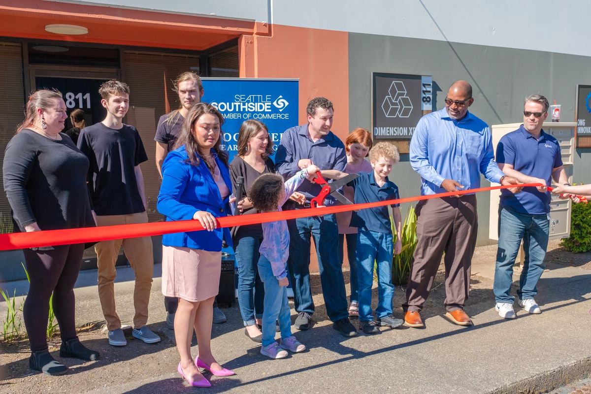 A crowd of adults and kids cuts a long red ribbon outside the 811 Industry Dr entrance, next to a Seattle Southside Chamber of Commerce banner and the Dimension XR sign.